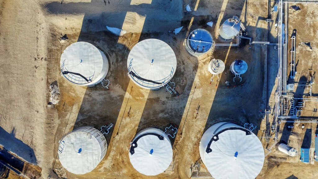 Aerial view of a large industrial storage tank facility with multiple white cylindrical tanks, connected pipelines, and processing units spread across a dry landscape under clear daylight.