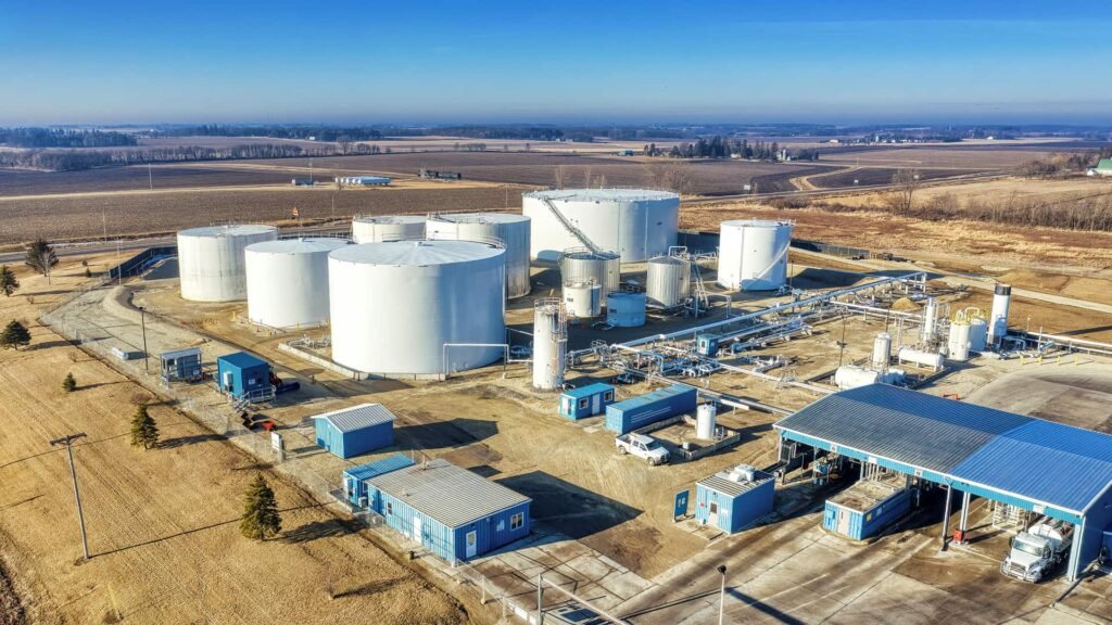 Top view of an industrial oil and fuel storage terminal featuring large circular storage tanks, metallic pipelines, and utility structures arranged in an organized layout on sandy terrain.