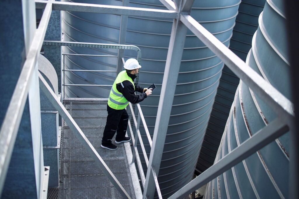 Worker inspecting large industrial storage tanks from a metal platform while using a tablet.